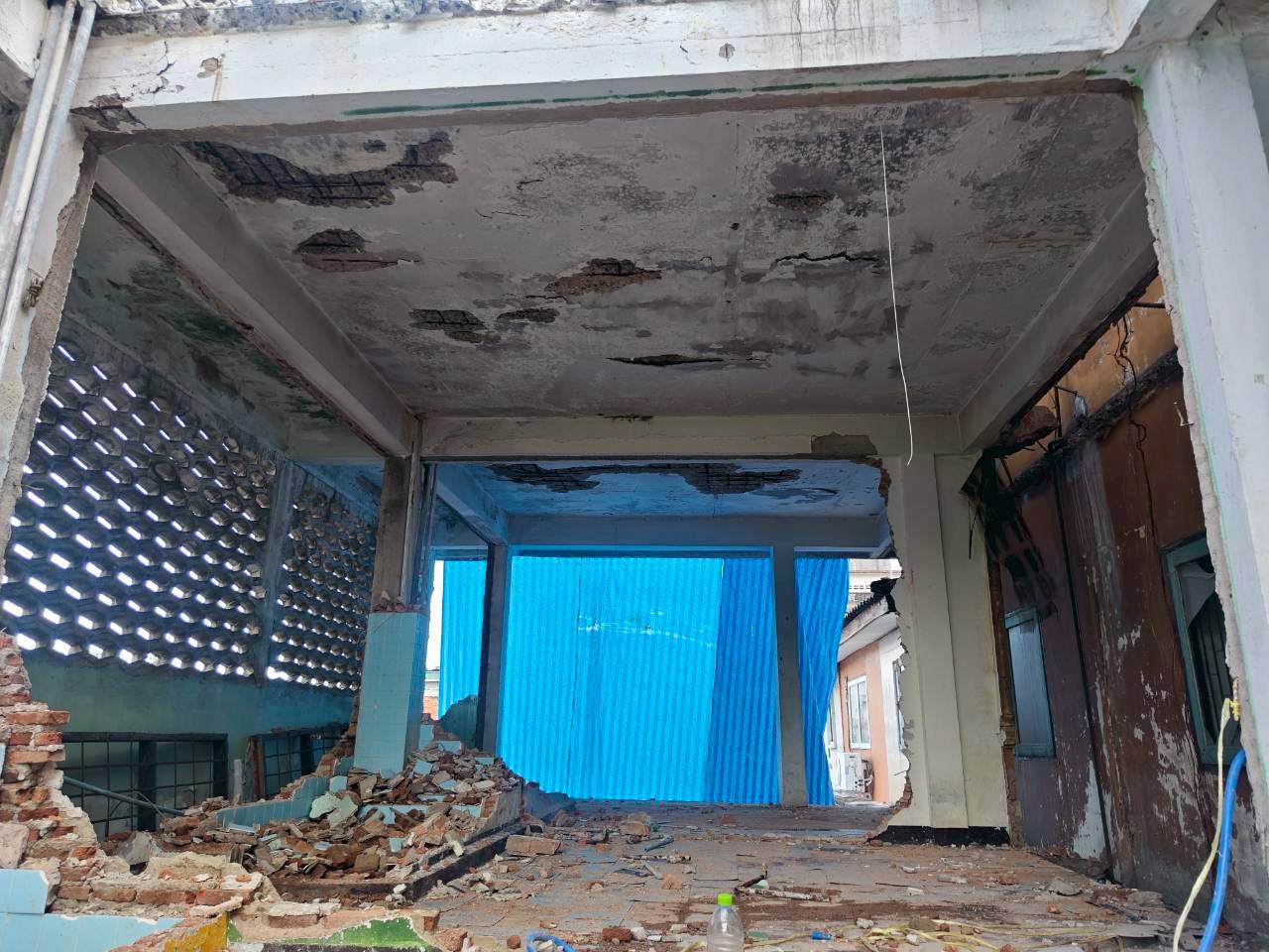 Ground-floor room at WITHIN during demolition with damaged ceiling, broken walls, and rubble piled beneath a blue temporary screen
